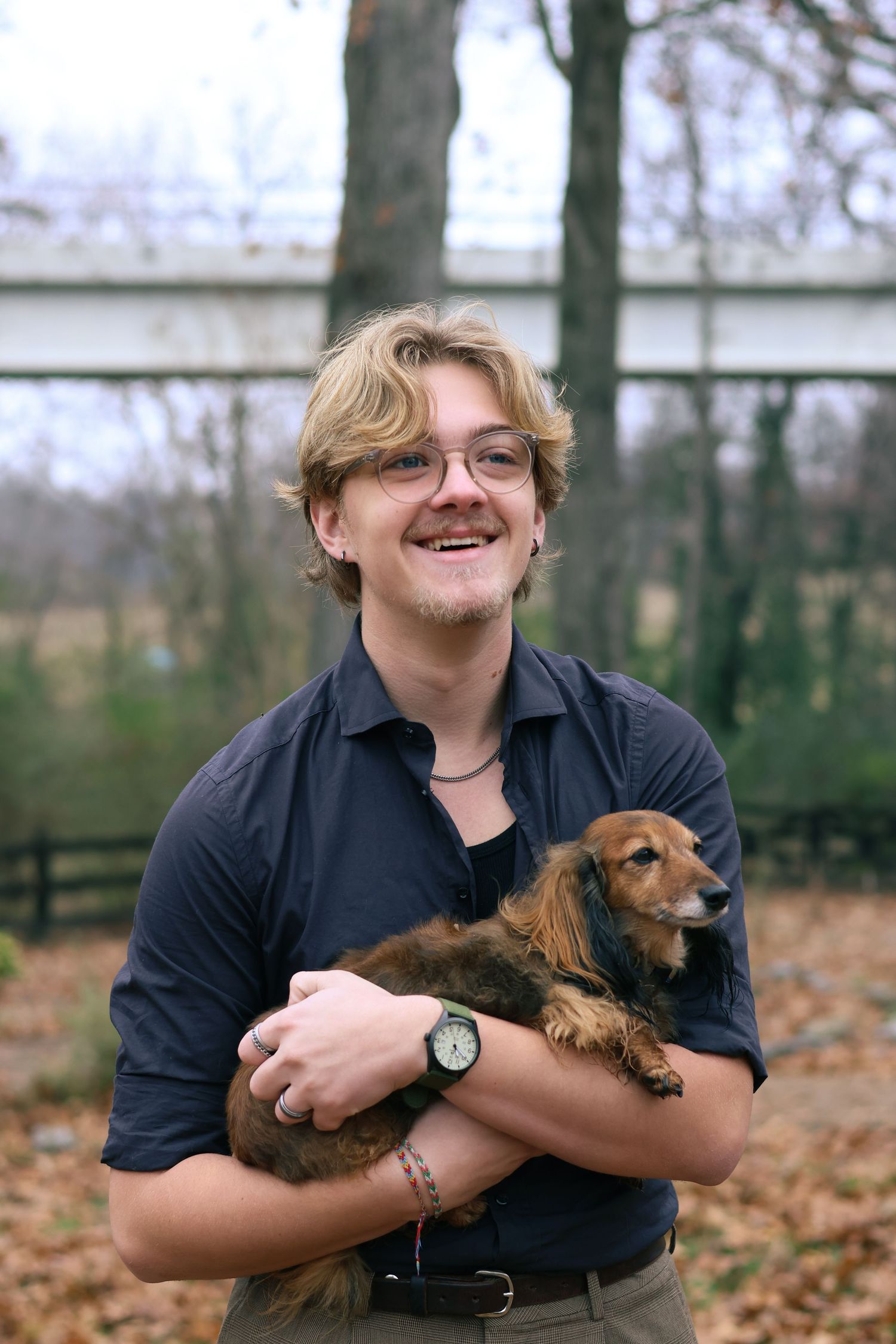 A person in a navy shirt holds and cuddles with a small brown dachshund dog while sitting among fallen autumn leaves.