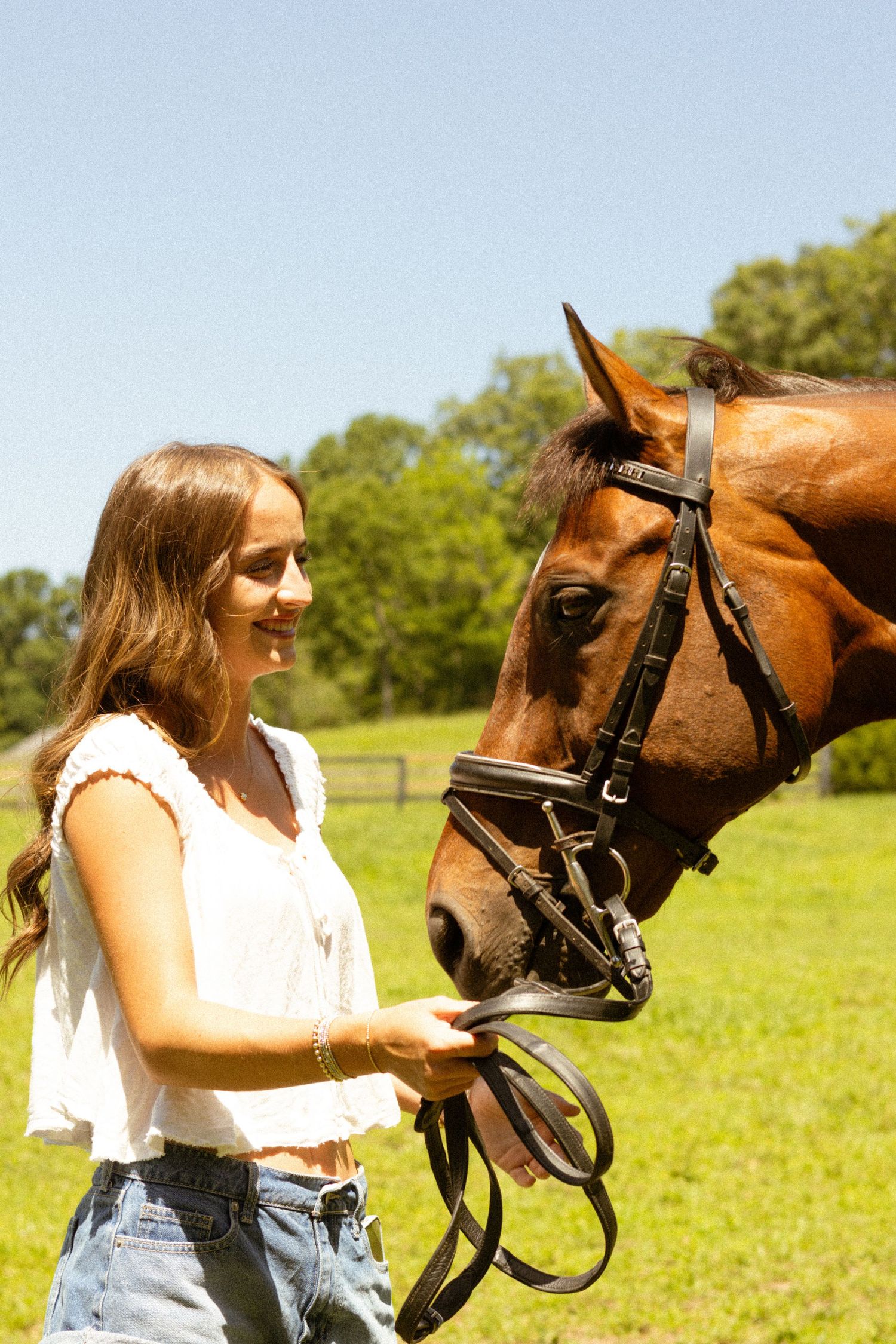 Young woman in white top and jeans smiling while holding reins of a brown horse outdoors.