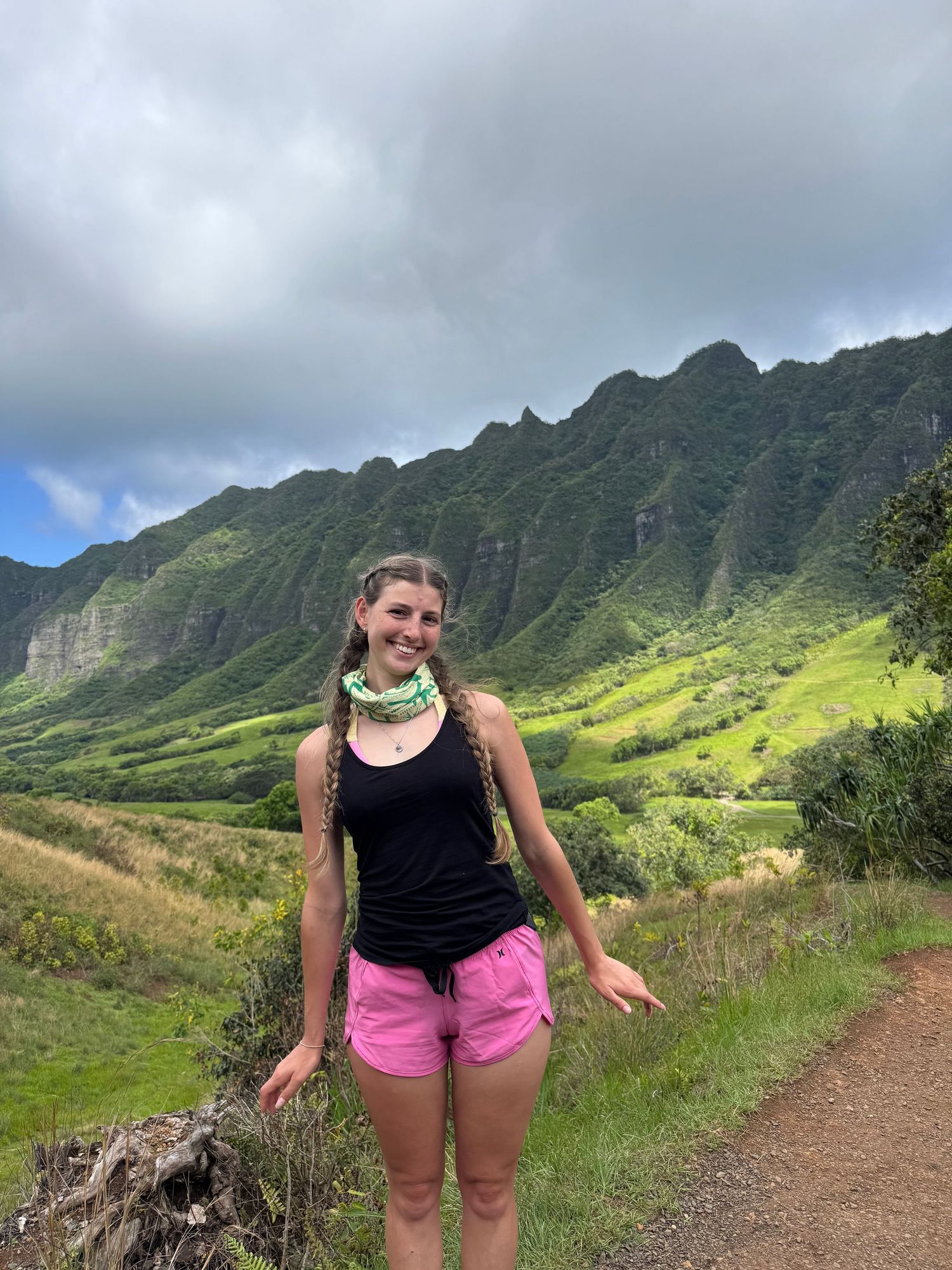Woman in pink shorts and black tank top poses on a hiking trail with lush green Koʻolau Mountains backdrop.