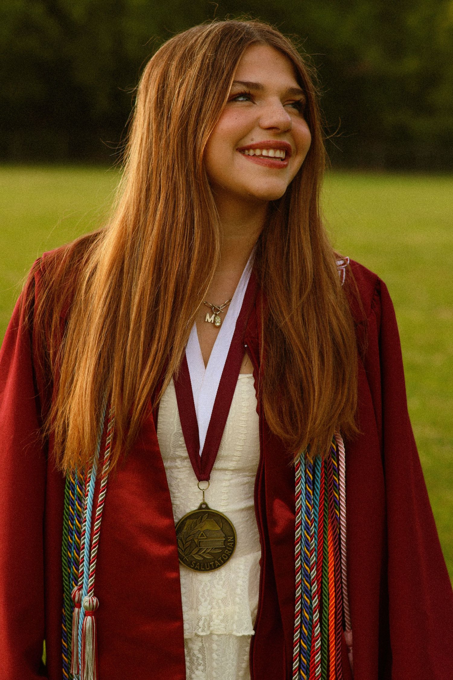 Smiling young woman in red graduation gown with colorful honor cords and medal, standing outdoors on green grass.