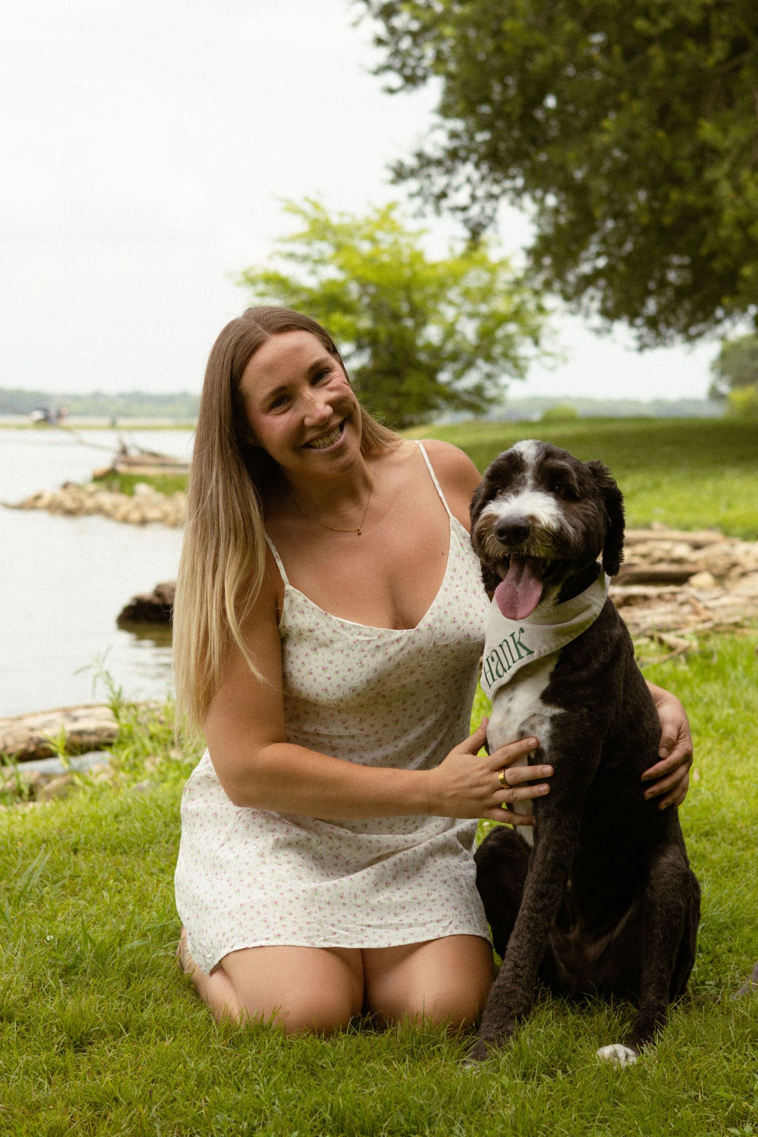Smiling woman in floral dress kneeling on grass with her black and white dog near a lake.