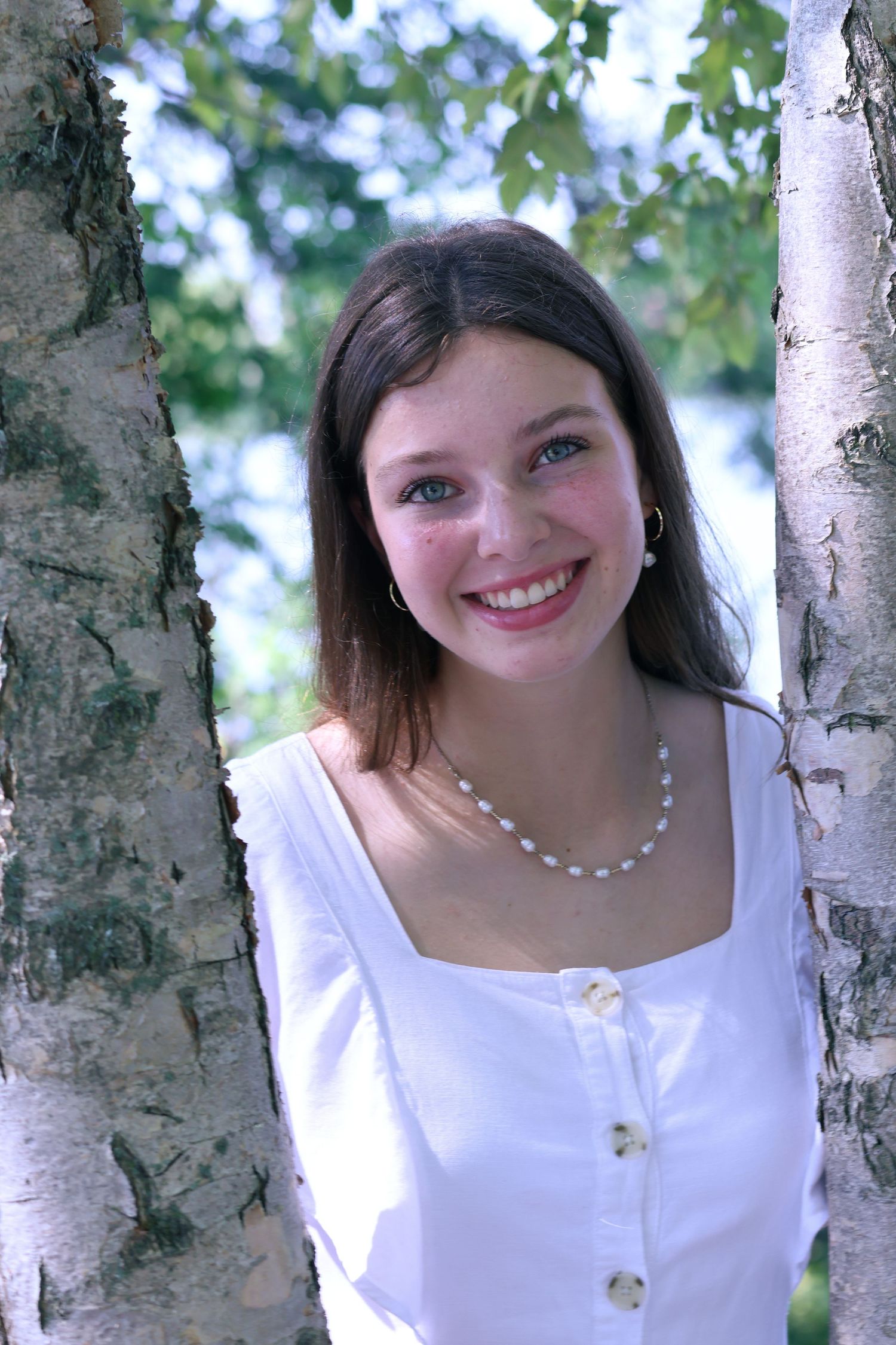Woman in white dress smiles between birch trees in natural outdoor setting.