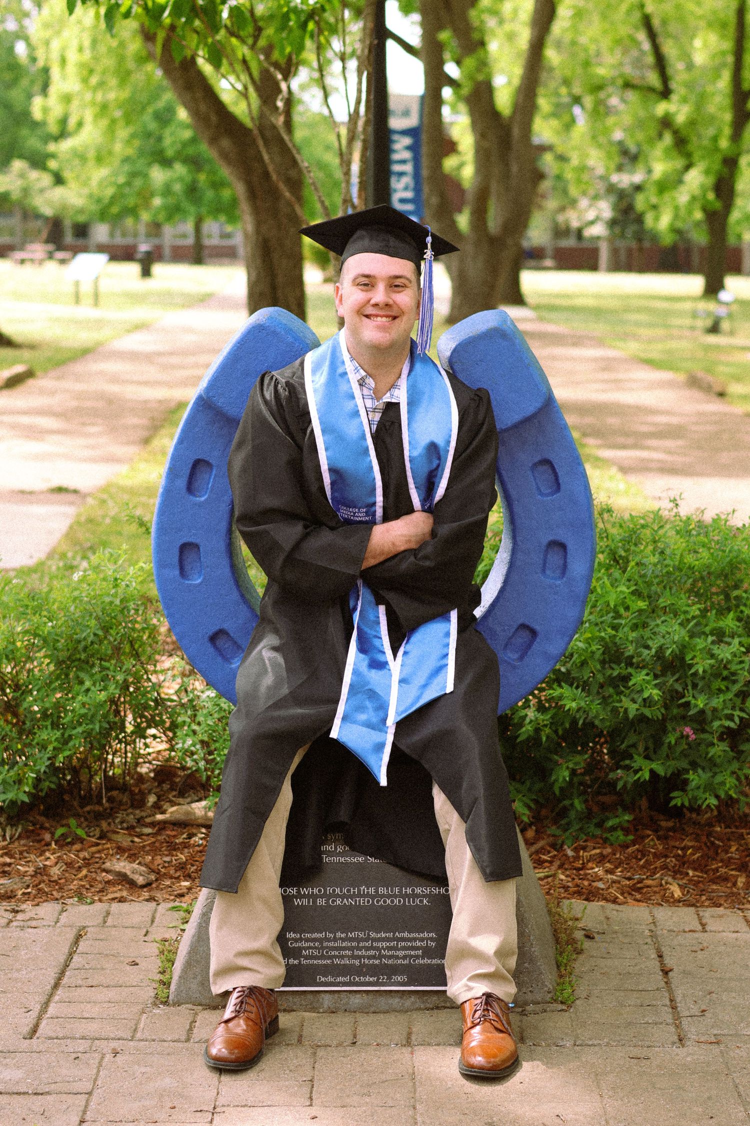 Graduate in cap and gown poses proudly in front of a large blue horseshoe statue on a tree-lined campus path.