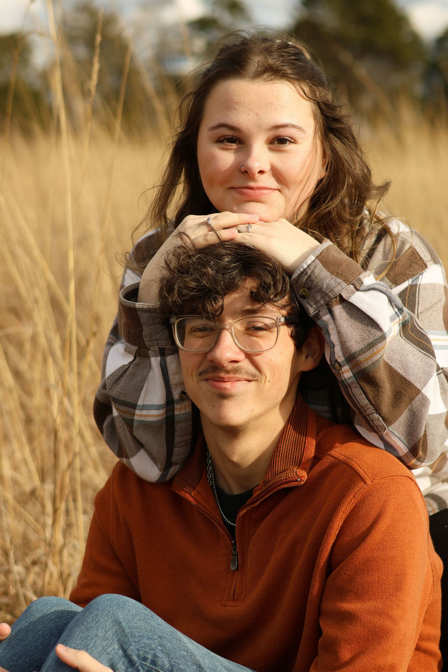 A person in a rust-colored sweater and glasses sits in a golden field during autumn.