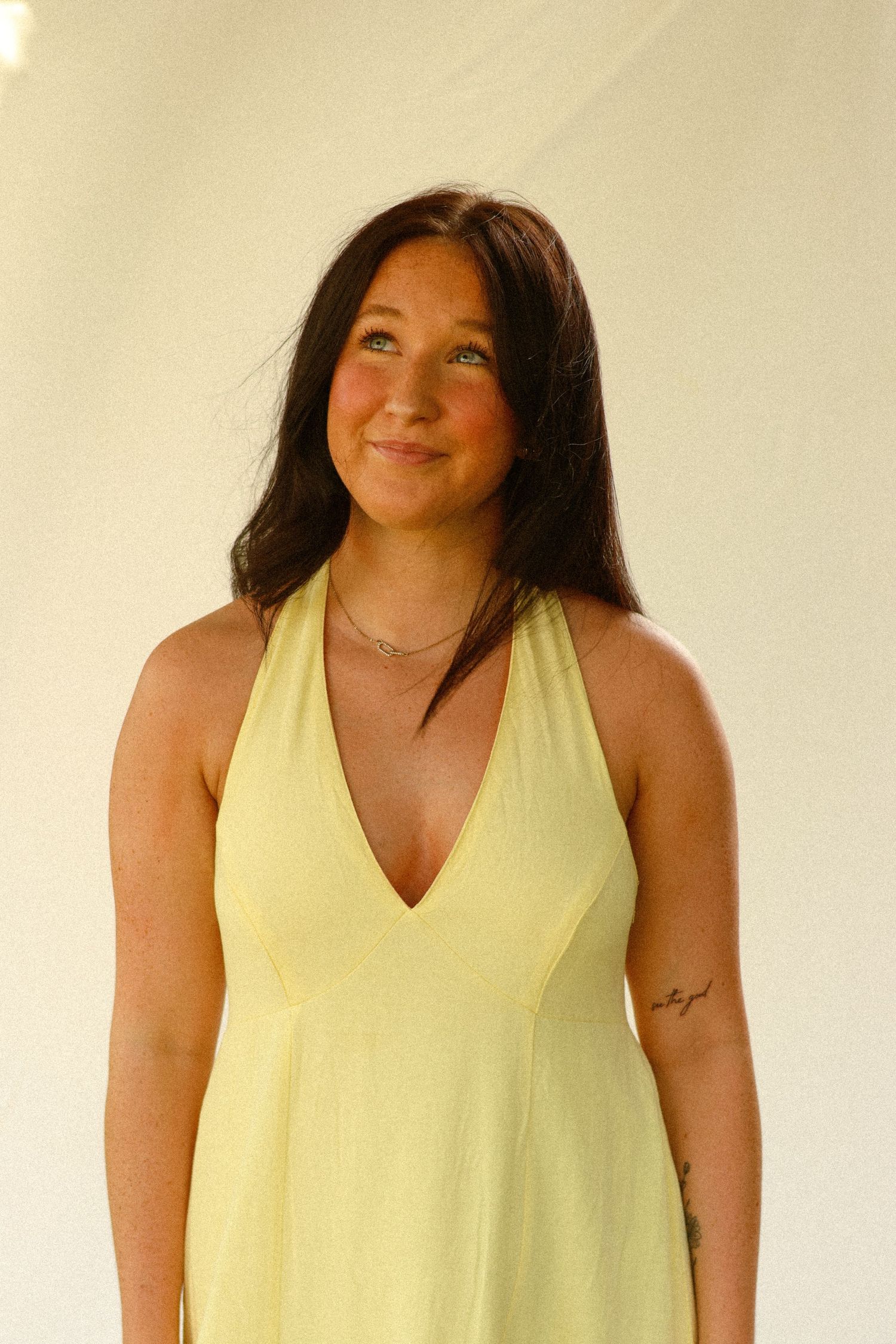 Young woman with dark hair smiling in a yellow halter dress with a delicate necklace and tattoo.