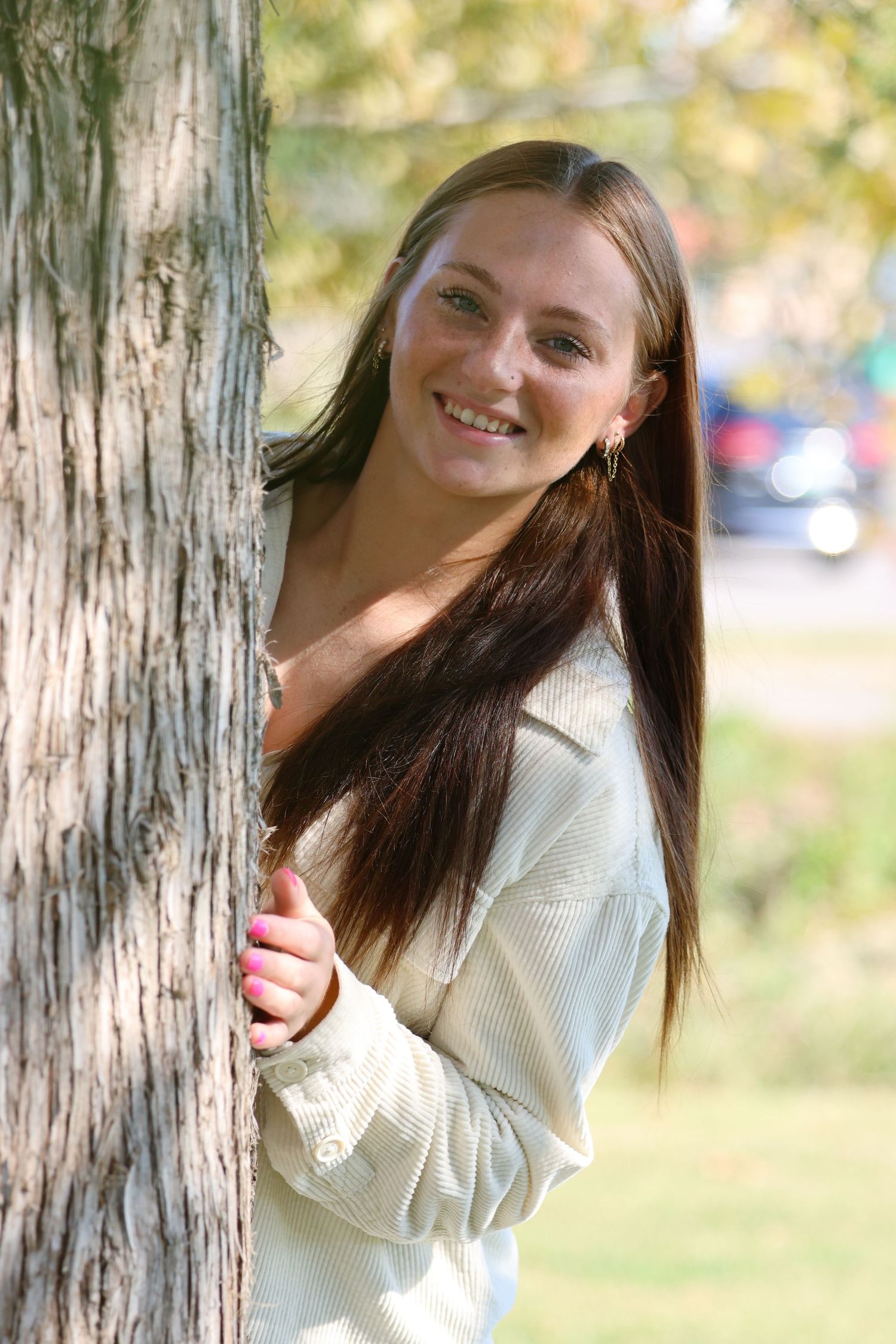 Person in white sweater posing playfully near tree trunks on a sunny day.