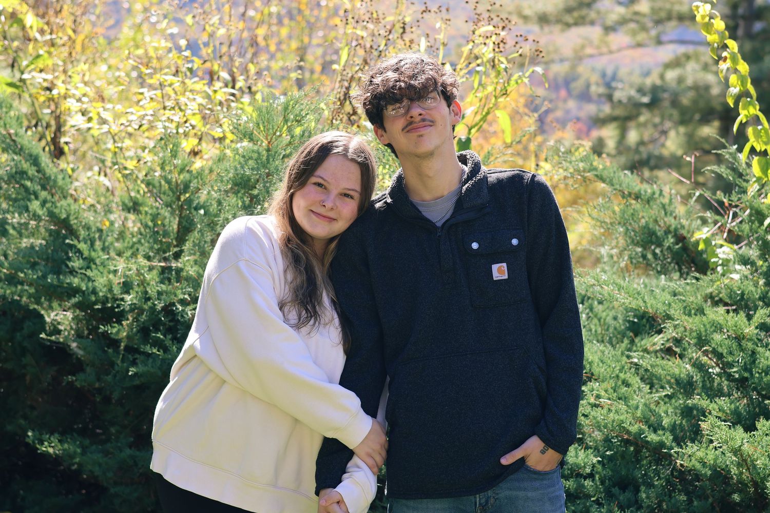 A playful couple in casual outfits enjoying a sunny autumn day in a wooded area.