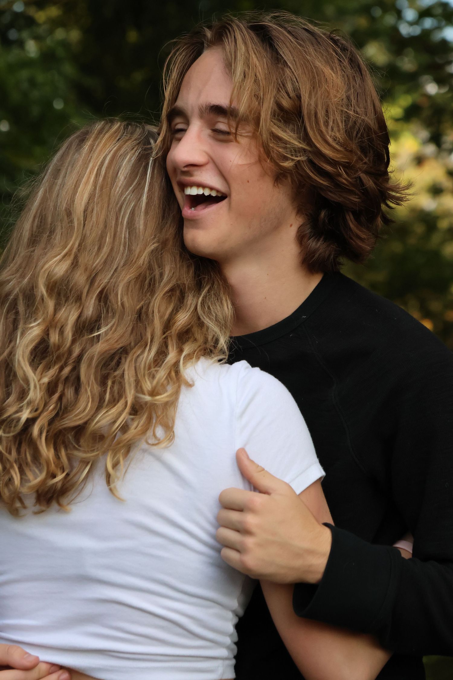 A joyful couple embraces and laughs together outdoors with trees in the background.