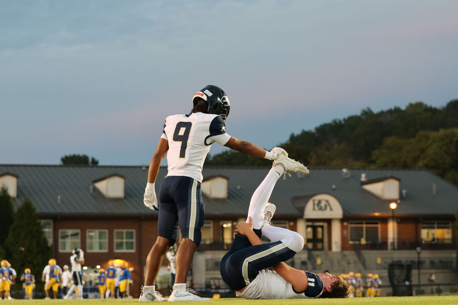 Football players and officials gather on a field during a game at sunset.