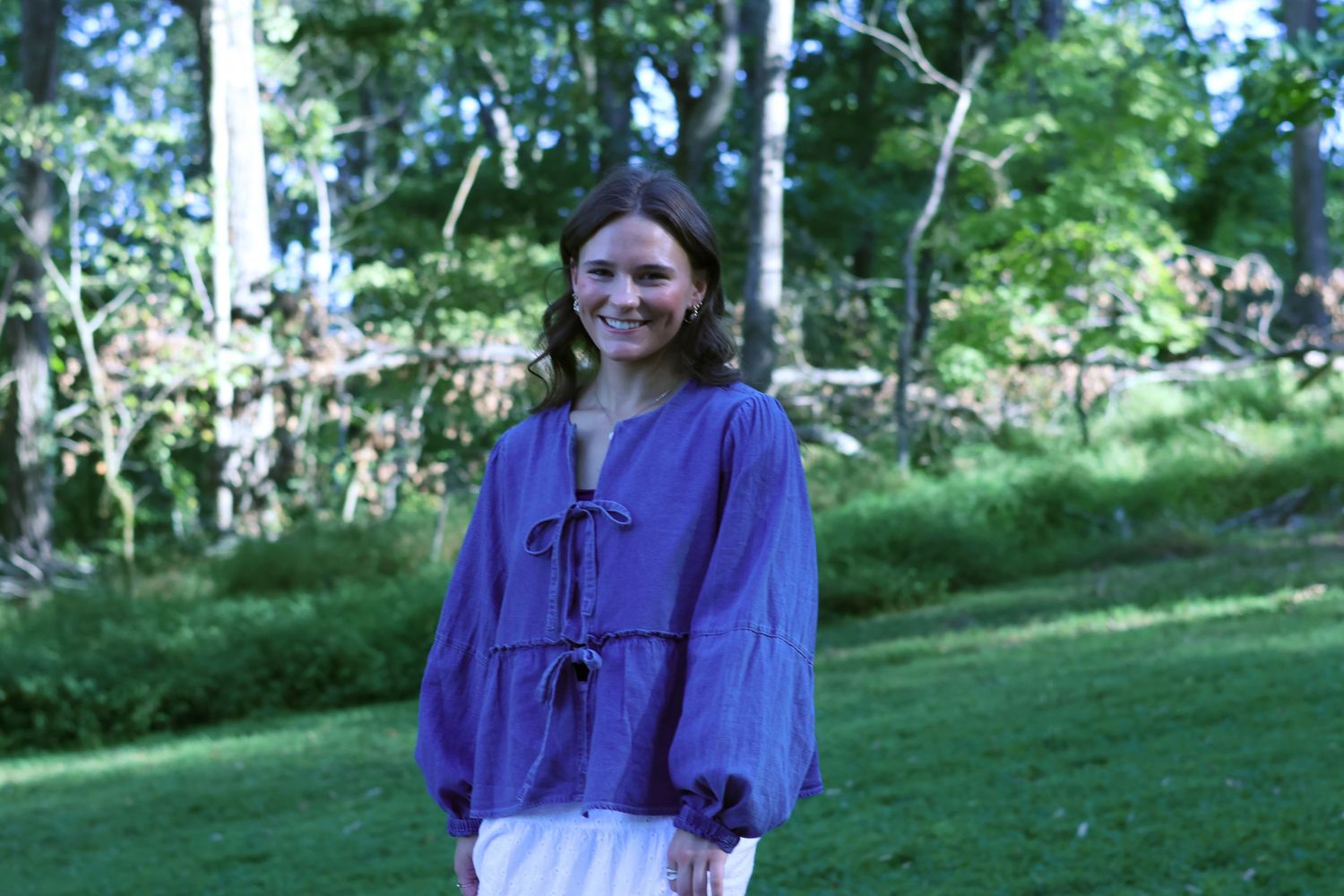 Woman in purple dress poses in sunlit wooded area.