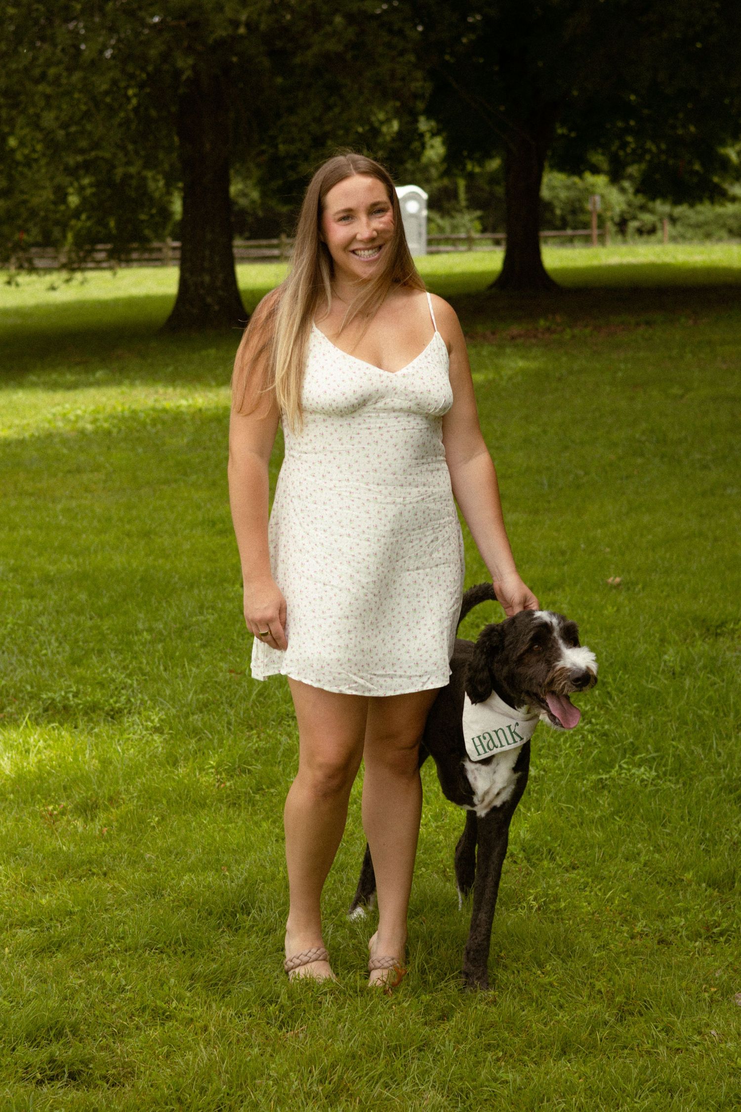 Smiling woman in white floral dress posing with a Great Dane dog on a green lawn surrounded by trees.