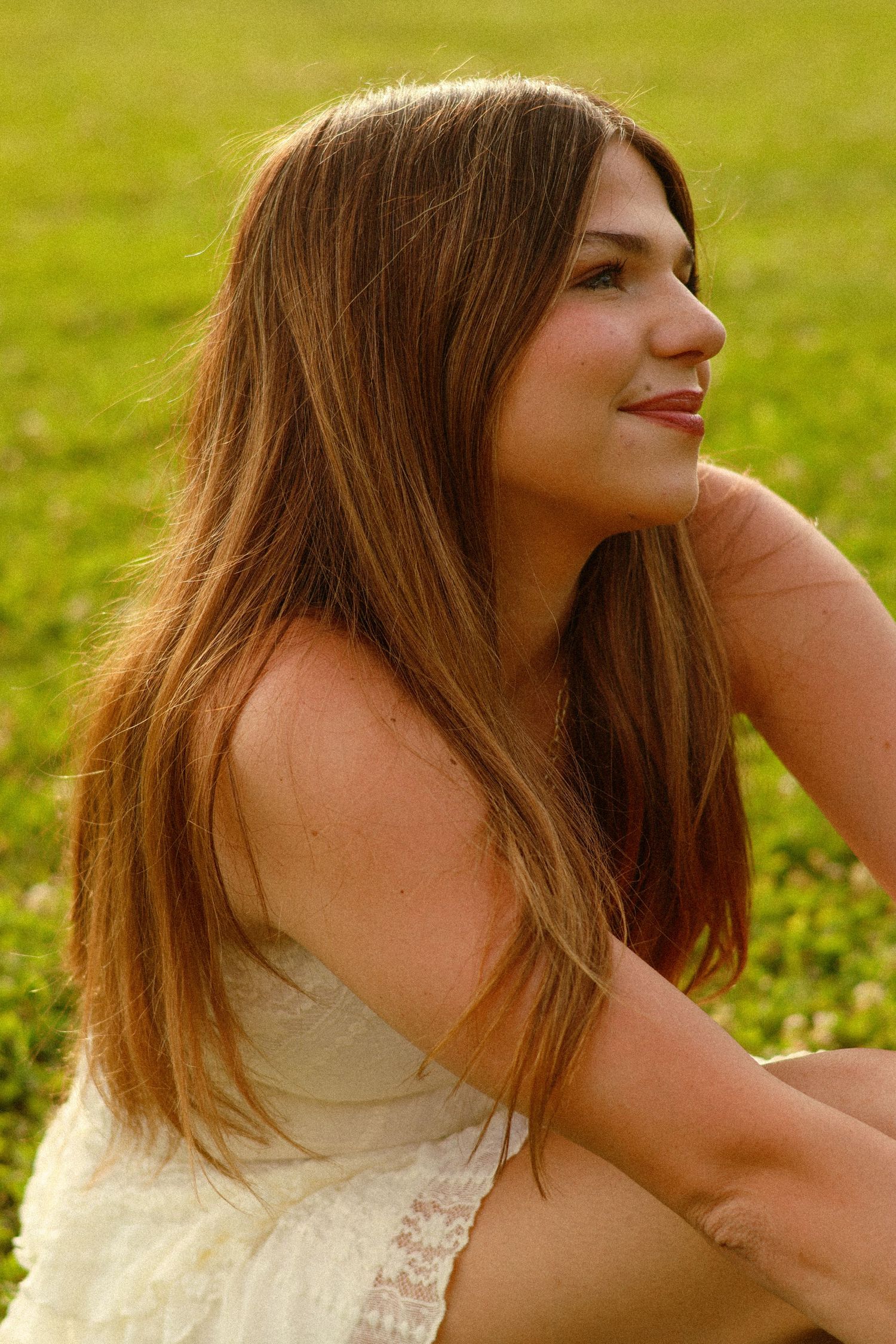 Young woman with long brown hair sitting in green grass field wearing white dress, smiling and looking away.