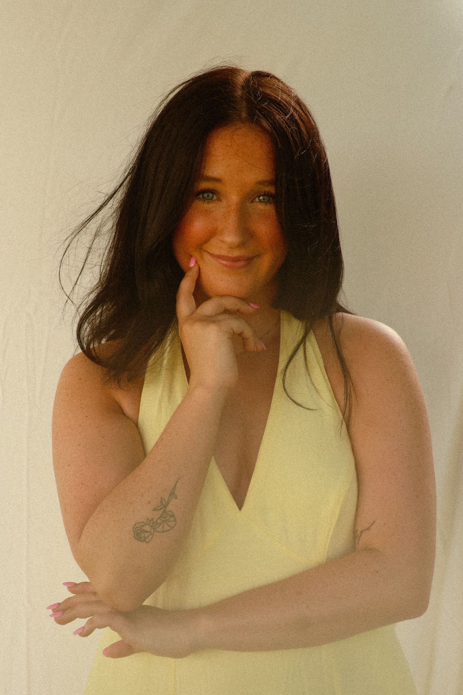 Young woman with dark hair in yellow dress smiling, hand on chin, tattoo on forearm, against white background.