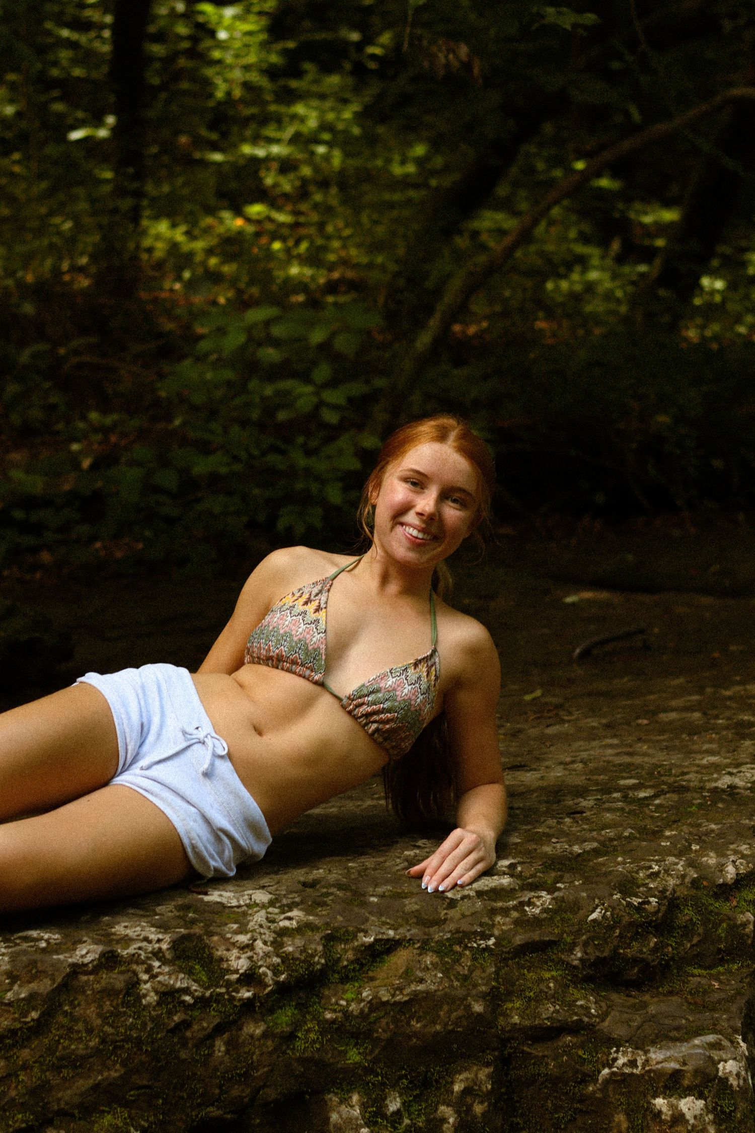 Smiling redhead woman in bikini top and white shorts relaxing on rocks in a lush forest setting.