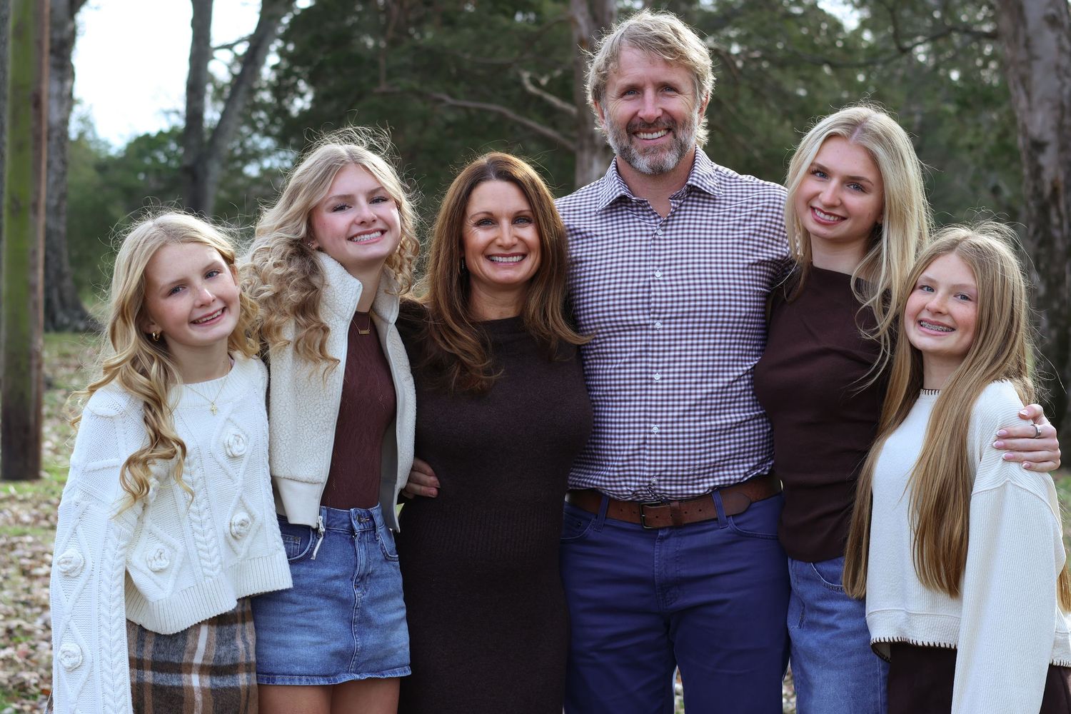 A family group poses together outdoors wearing coordinated casual outfits against a natural wooded backdrop.