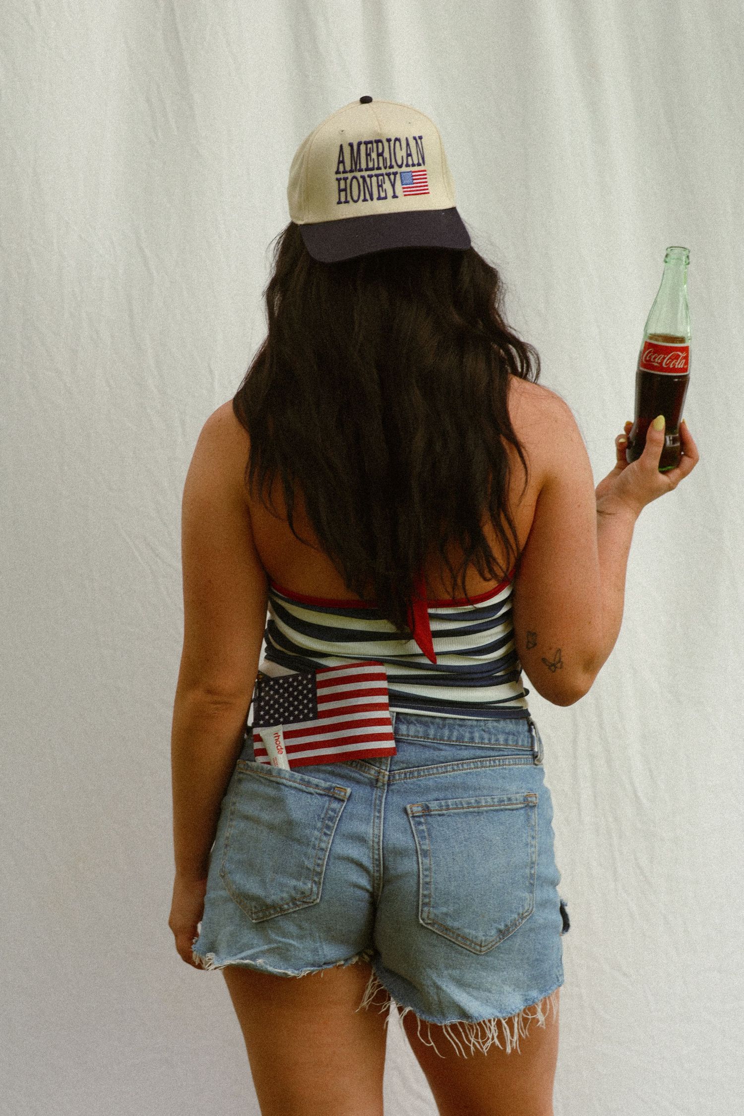Woman in denim shorts holding Coca-Cola bottle, wearing American Honey trucker hat and American flag tucked in pocket.