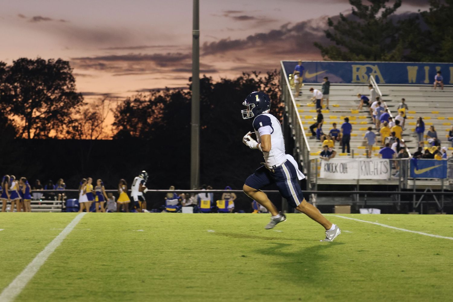 Football players compete on a field against a dramatic sunset sky backdrop.