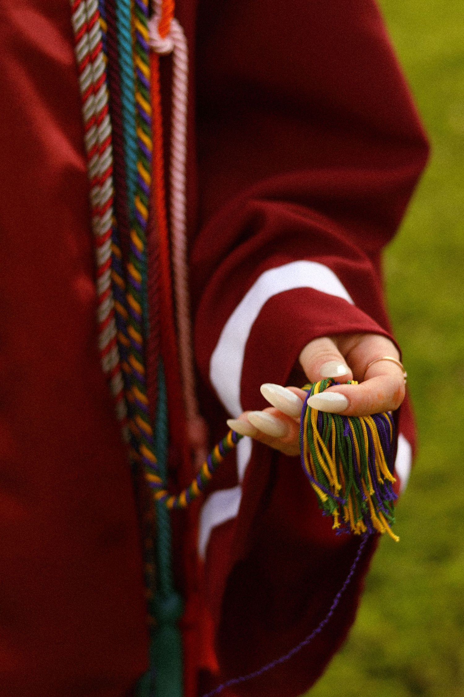 Graduate in red gown holds colorful tassel with braided honor cords draped over shoulder at graduation ceremony.