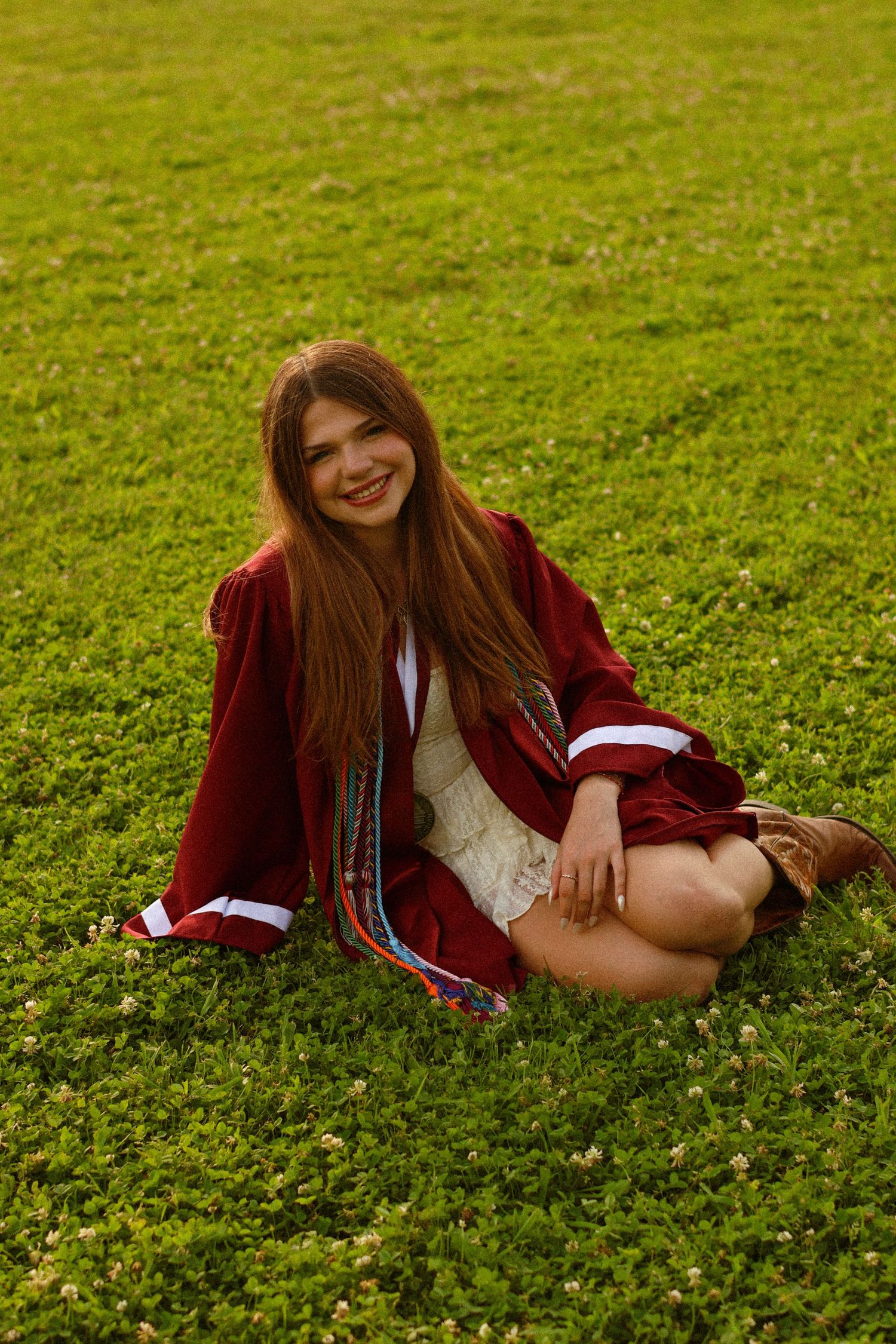 Happy young woman in red graduation robe sitting cross-legged on lush green grass smiling at camera.