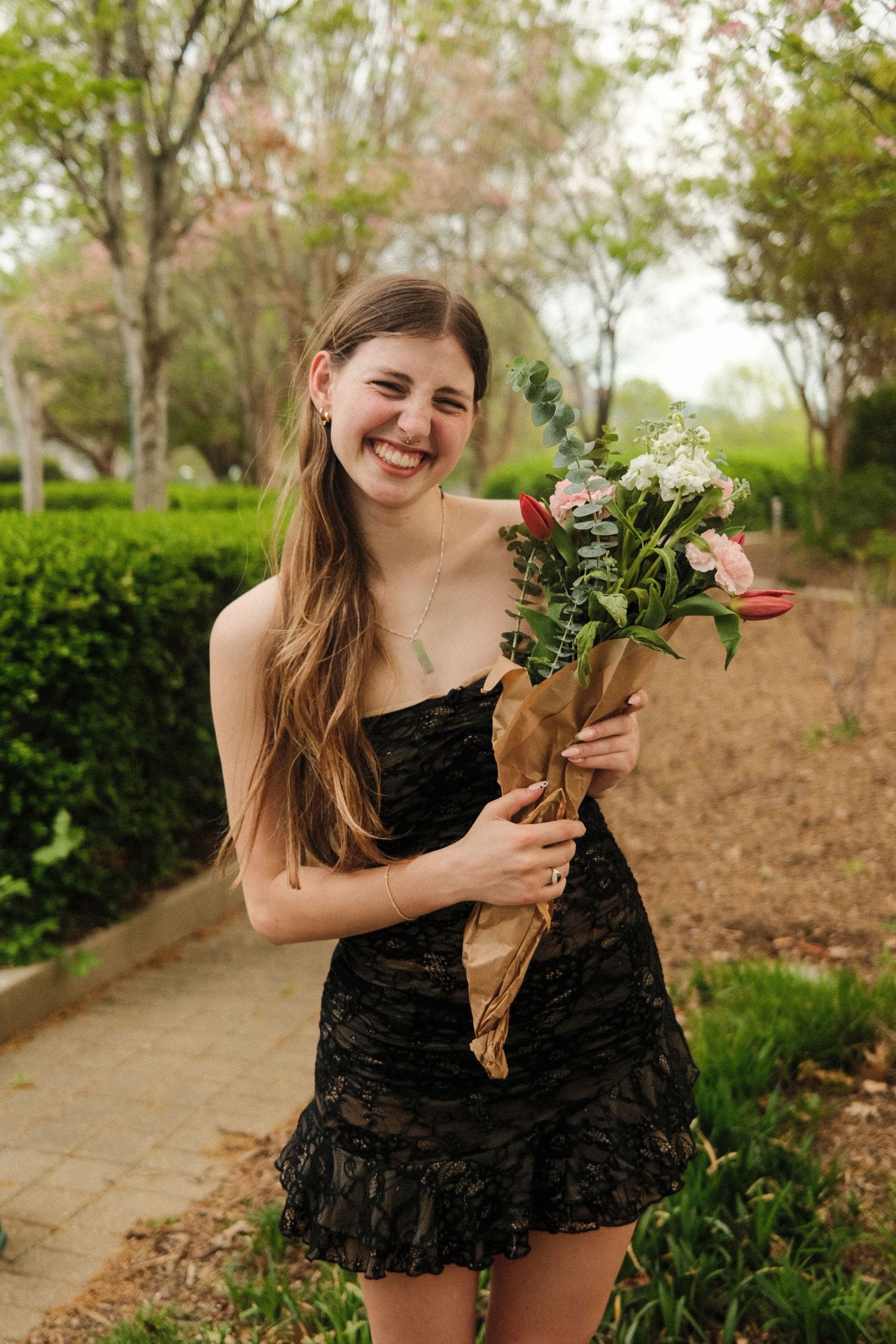 A joyful moment captured outdoors as someone holds a wrapped bouquet of flowers while wearing a black dress.