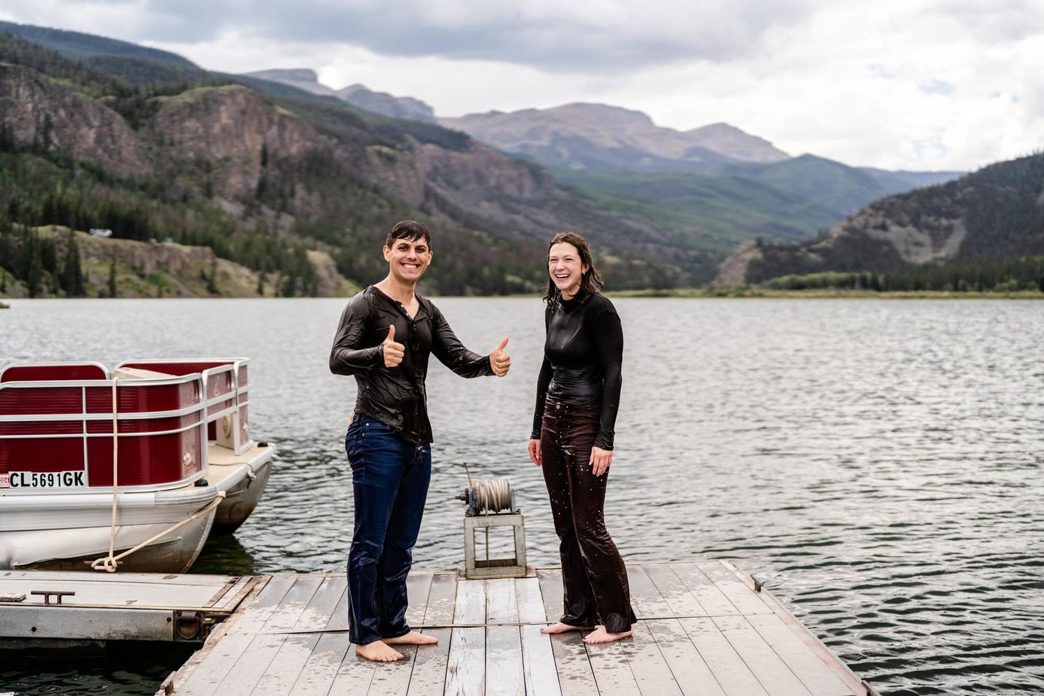 Two people standing on a wooden dock by a mountain lake surrounded by scenic views.