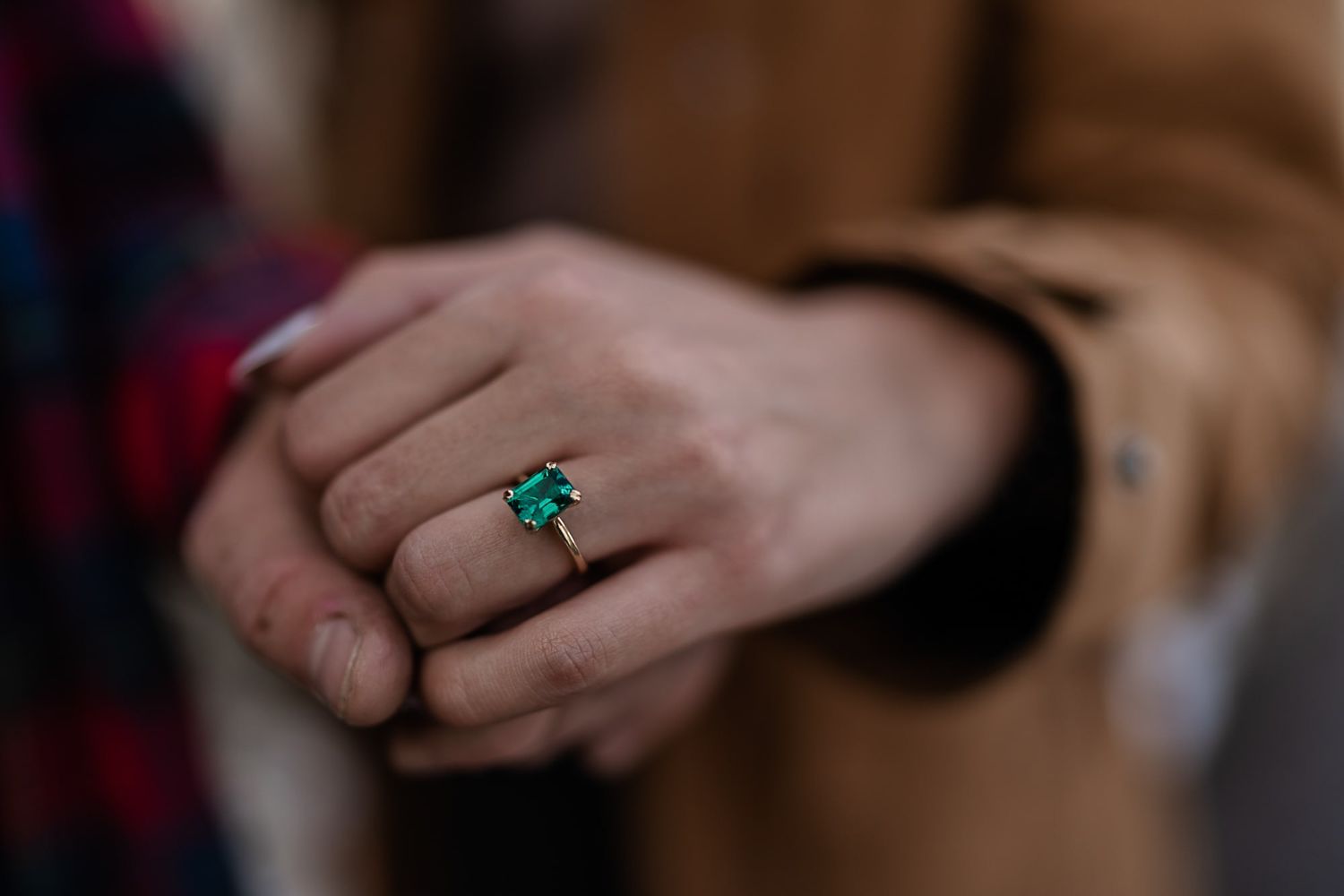 Close-up of an emerald engagement ring worn on a hand against a brown sleeve.