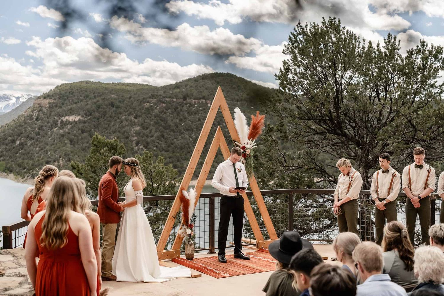 A mountain wedding ceremony takes place under a wooden triangle arch overlooking a scenic lake and hills.