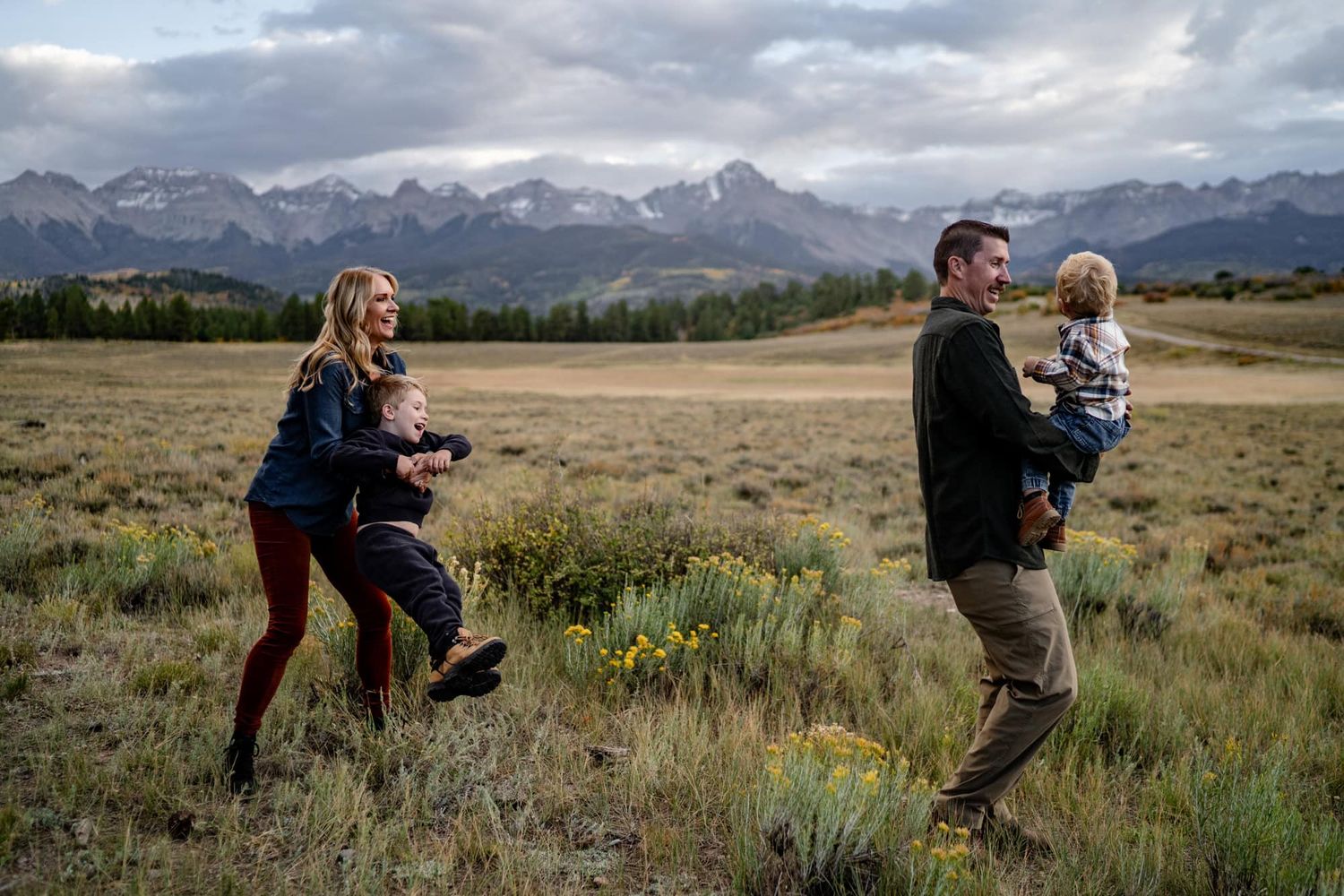 Family plays together in a meadow with dramatic peaks and cloudy skies in the background. Captured by a photographer near Ridgway Colorado.