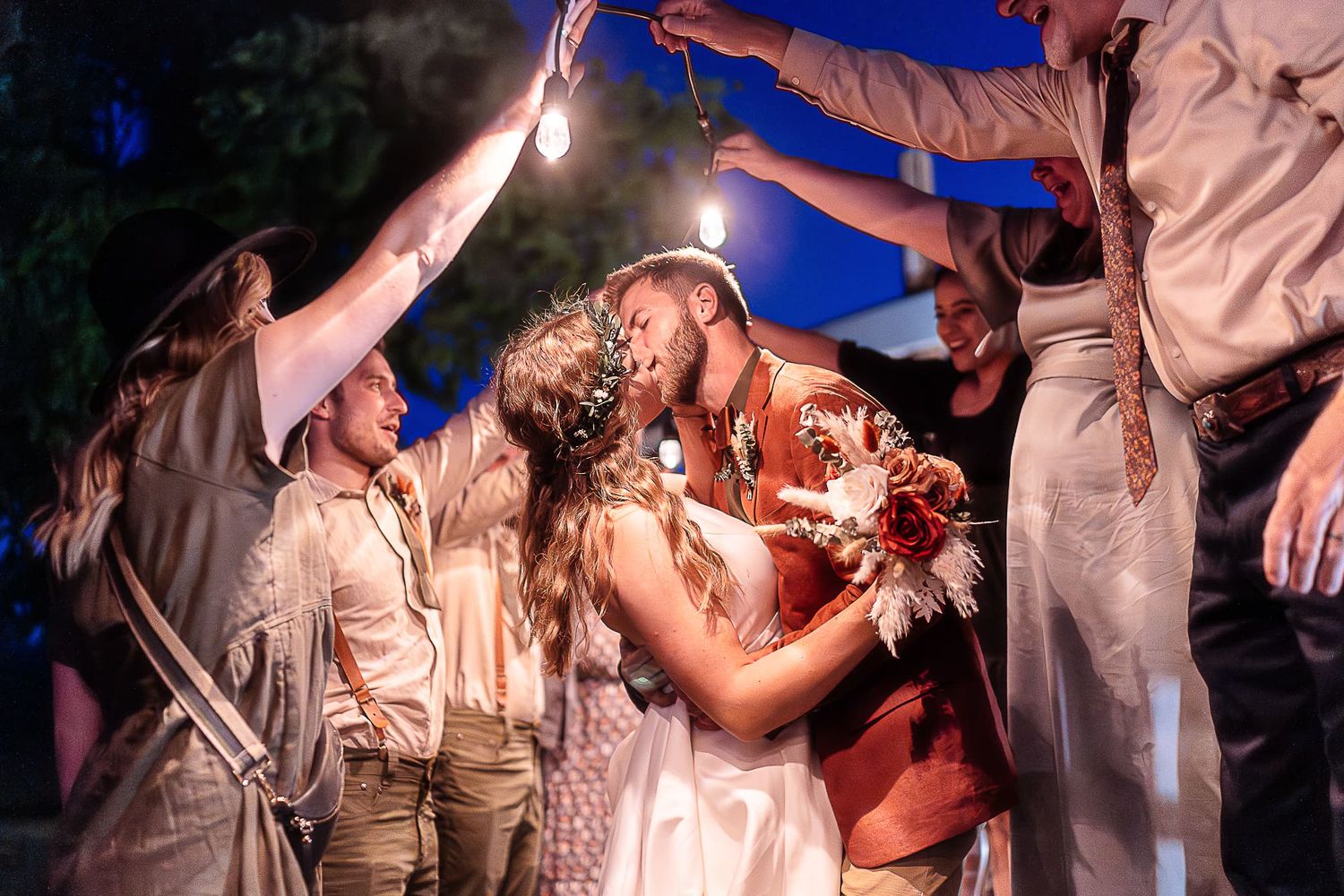 Newlyweds share romantic kiss under sparklers held by wedding guests at night.
