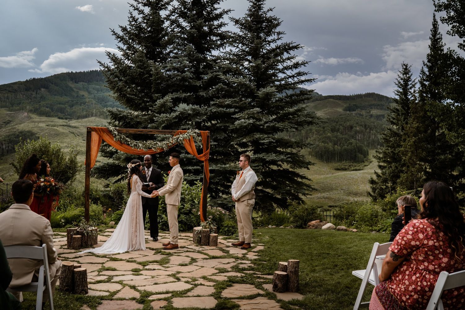 Mountain wedding ceremony under wooden arch with evergreen trees and dramatic cloudy sky backdrop.