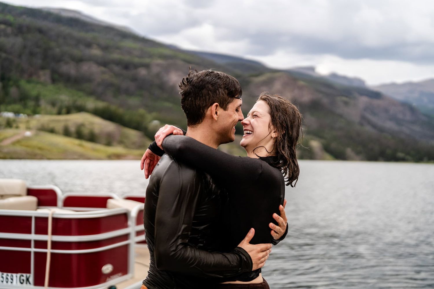 Romantic embrace on a dock with a mountain lake and pontoon boat in the background.