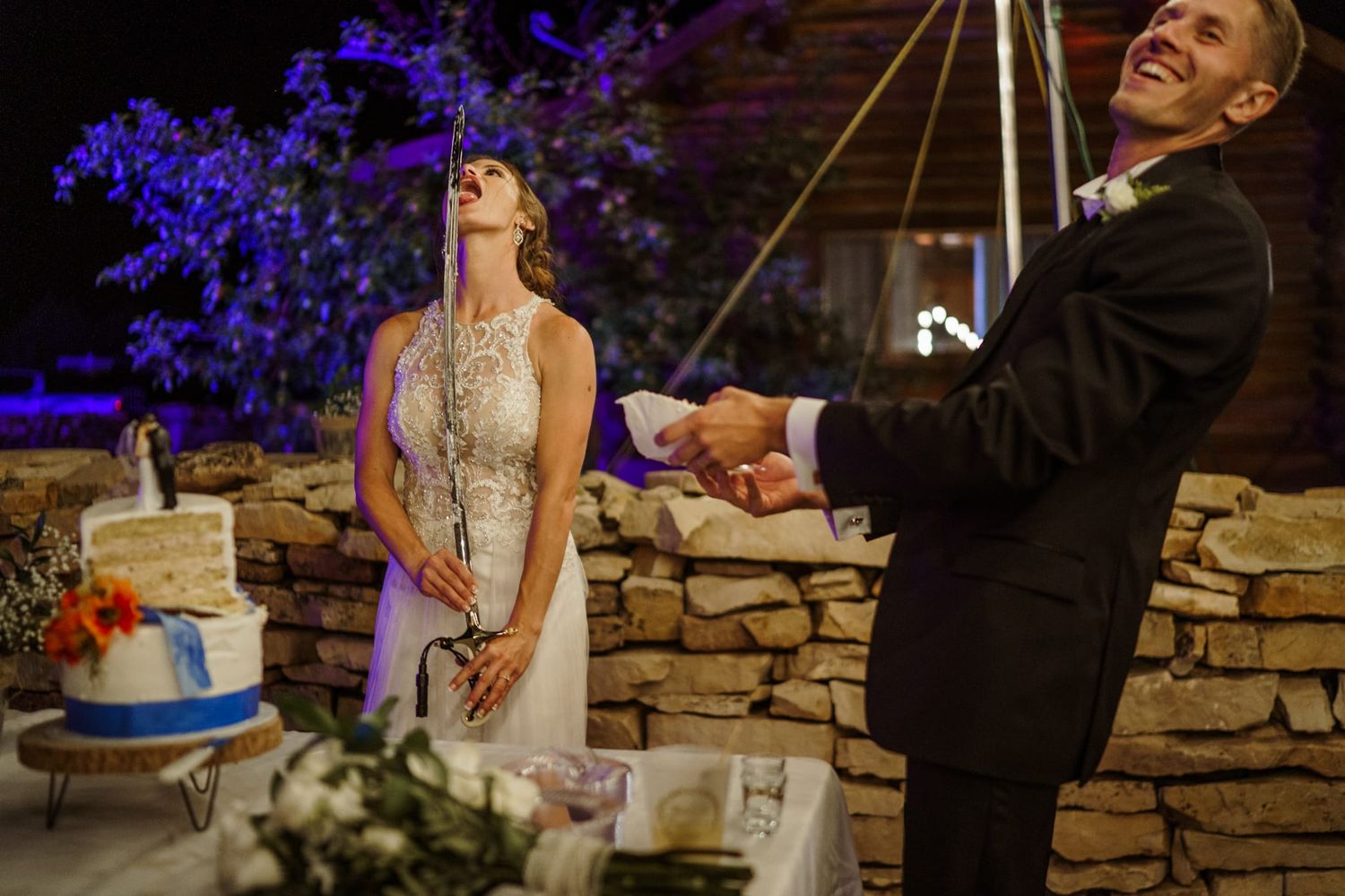 Wedding cake cutting moment at an outdoor evening reception with blue lighting.