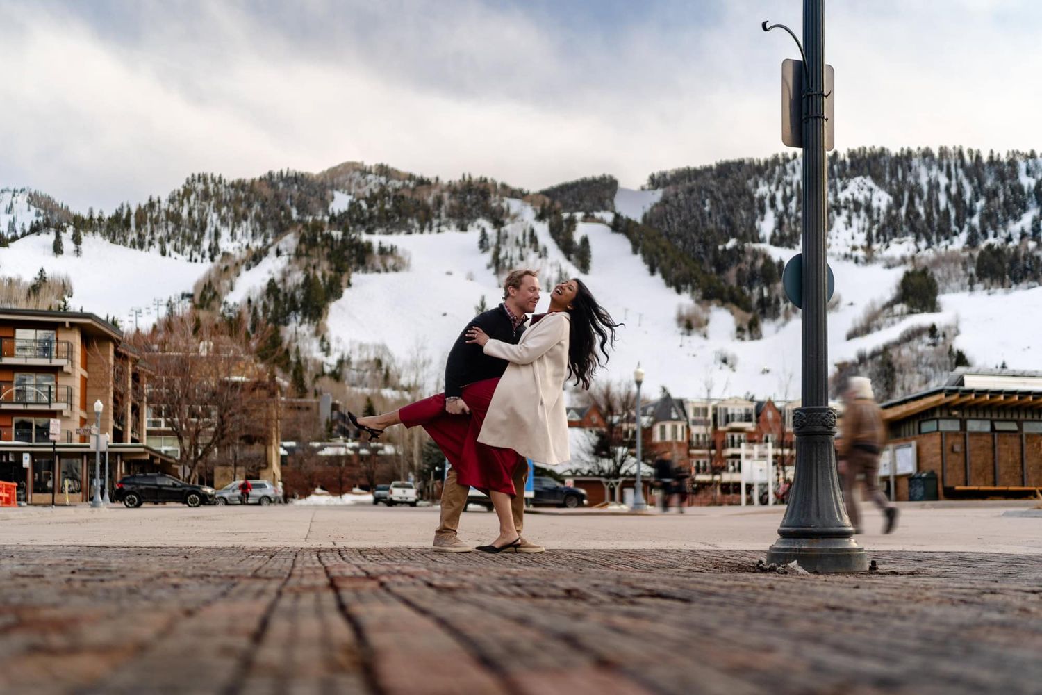 A couple dancing joyfully in the snow with a mountain ski resort in the background.