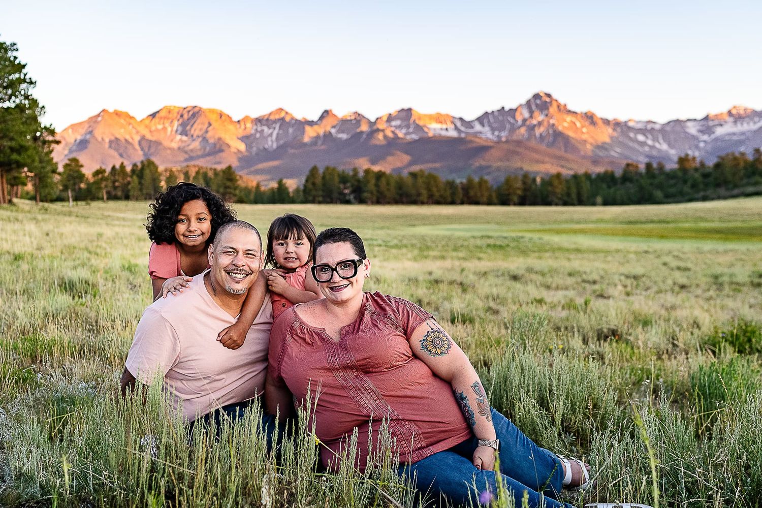 Family group sitting together in meadow with mountain range backdrop at sunset.