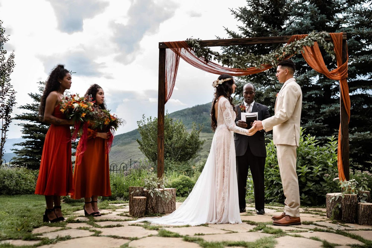 A small outdoor wedding ceremony takes place under a wooden arch with mountain views and evergreen trees in the background.