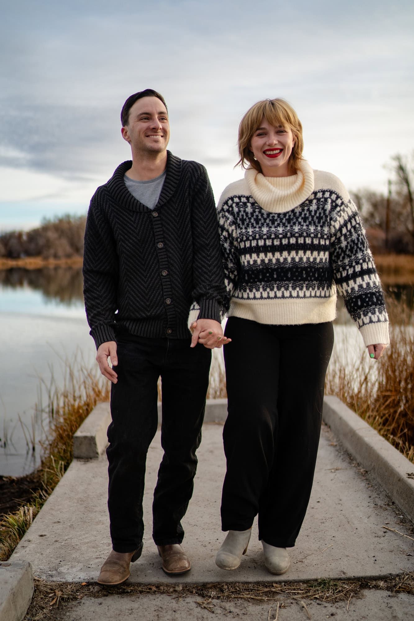 A couple walks hand in hand on a path by a lake, wearing cozy winter sweaters.