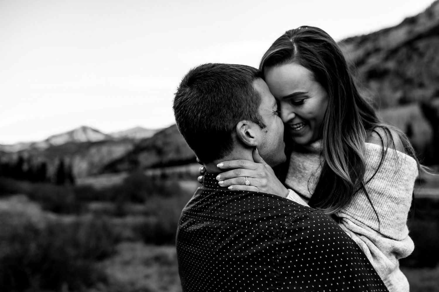 An intimate black and white portrait of a couple embracing against a mountain backdrop.