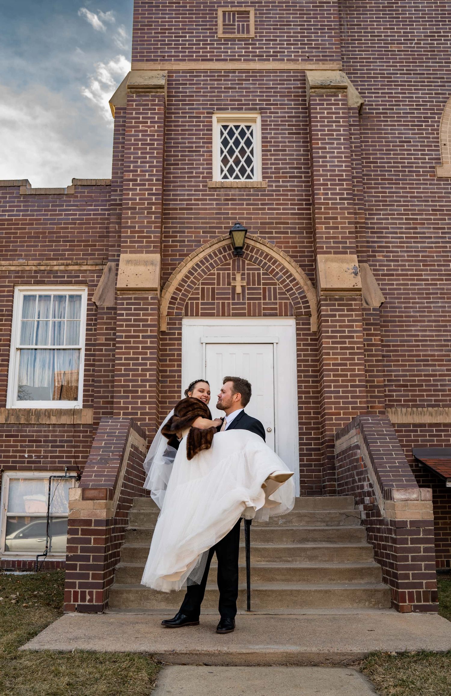 Groom carries bride in white dress in front of brick church entrance.