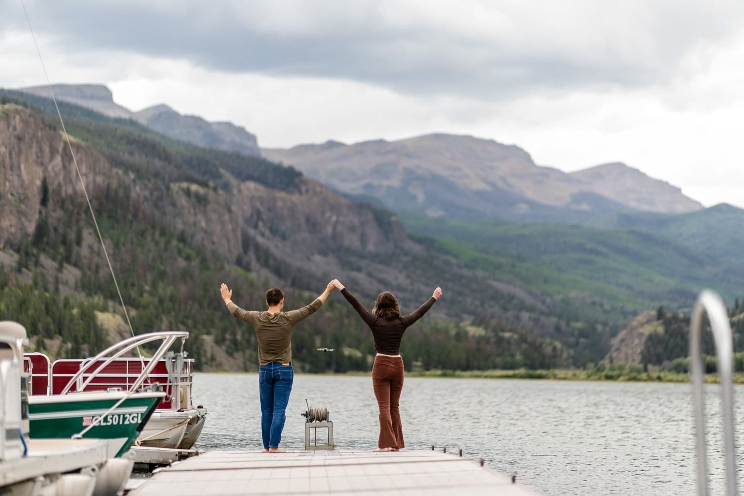 Two people stand with arms raised on a dock overlooking a mountain lake with dramatic cliffs in the background.