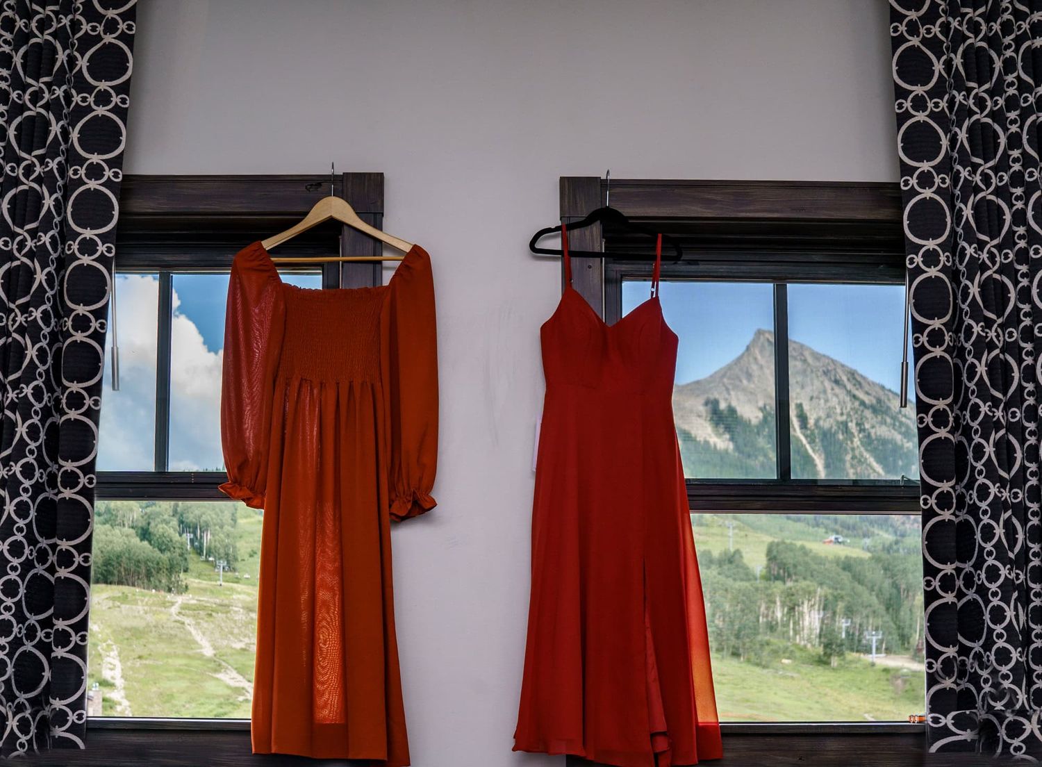 Two red dresses hang on hangers against white wall with mountain views through windows in background.