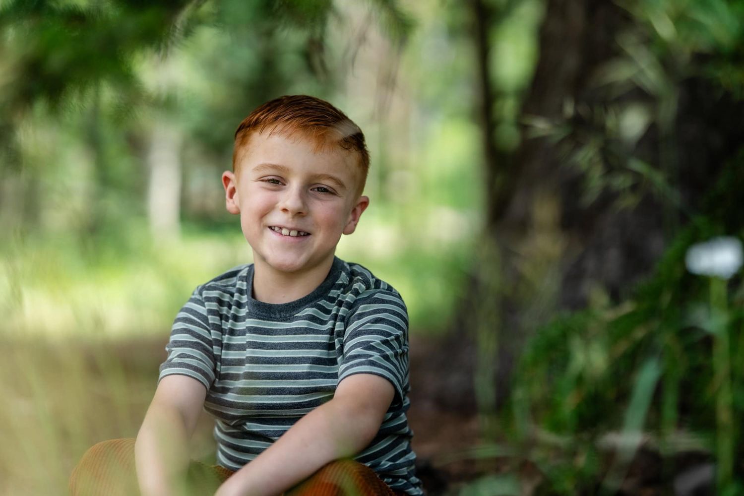 Young man in striped shirt smiling brightly in natural outdoor setting.