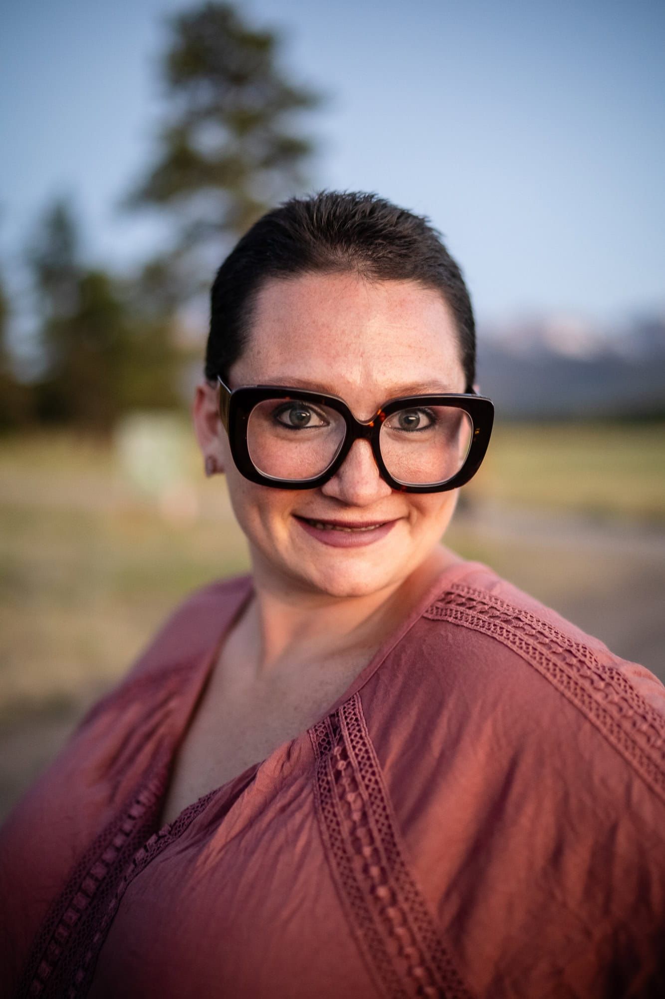 A person wearing glasses and a mauve sweater smiles warmly at the camera outdoors.
