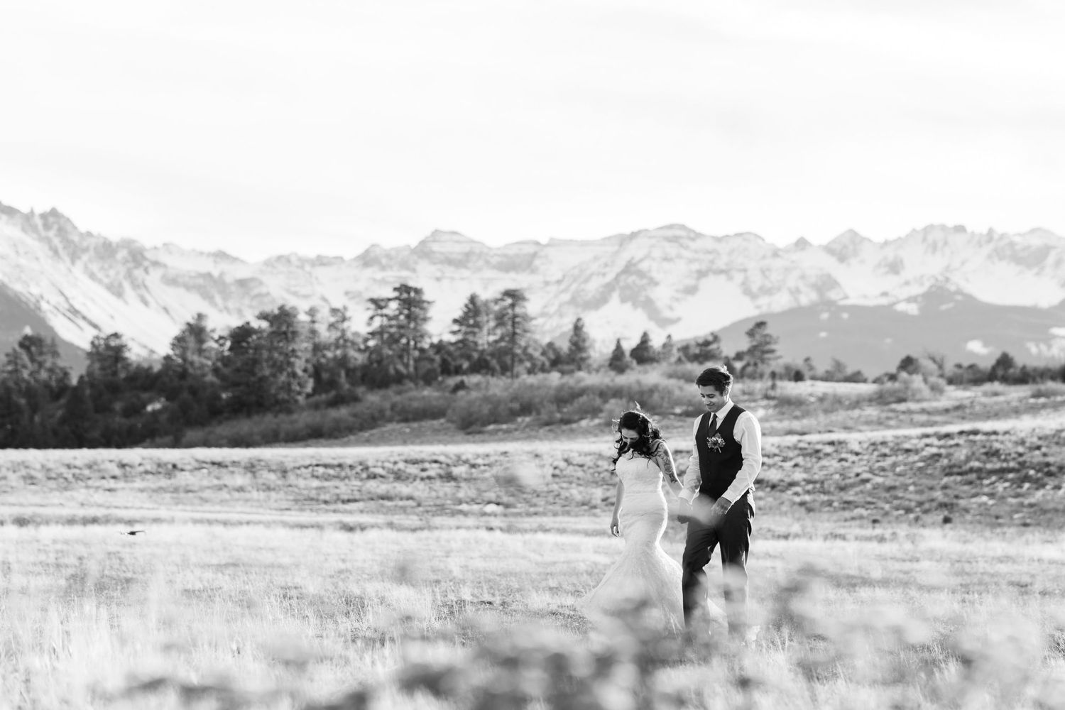 A romantic couple walks hand in hand through a meadow with snow-capped peaks in the background.
