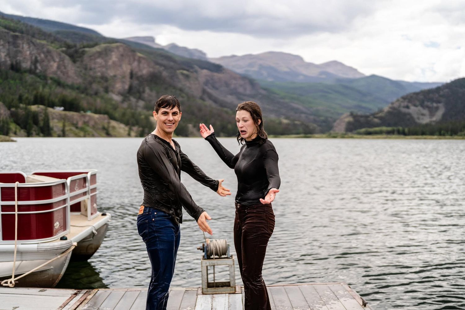 Two people sharing a fun moment on a wooden dock with mountains and lake in the background.