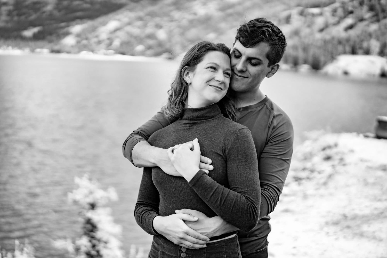 A couple shares an intimate embrace while standing by a scenic lake in black and white.