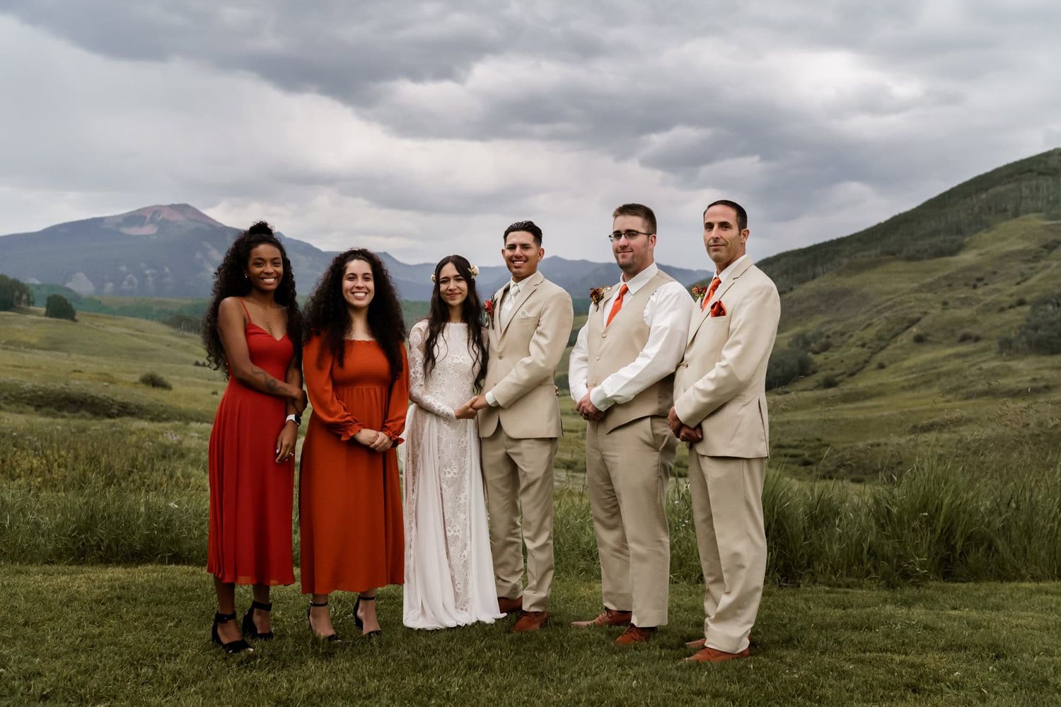 Wedding party in formal attire poses together against backdrop of rolling green hills and dramatic cloudy sky.
