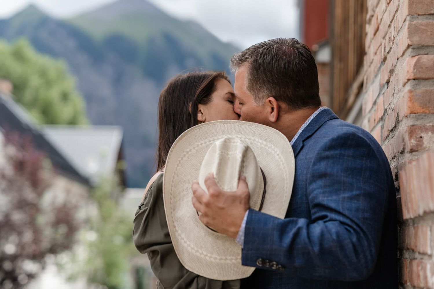 Romantic kiss shared behind white cowboy hat with mountain scenery.
