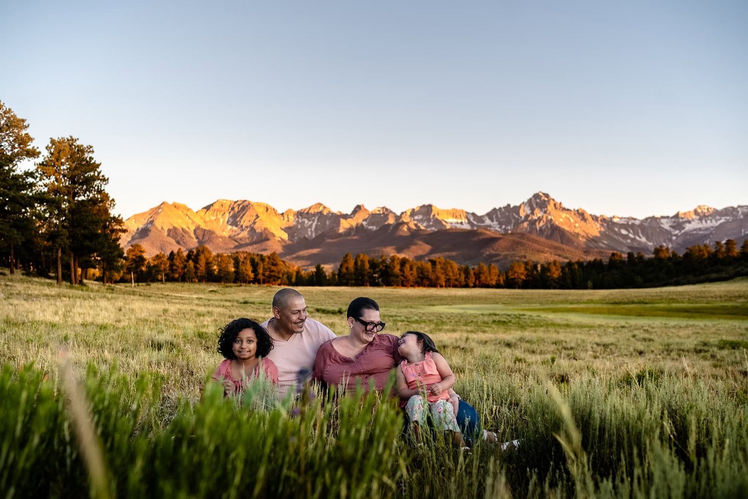 A family sits together in tall grass with mountain peaks glowing at sunset.