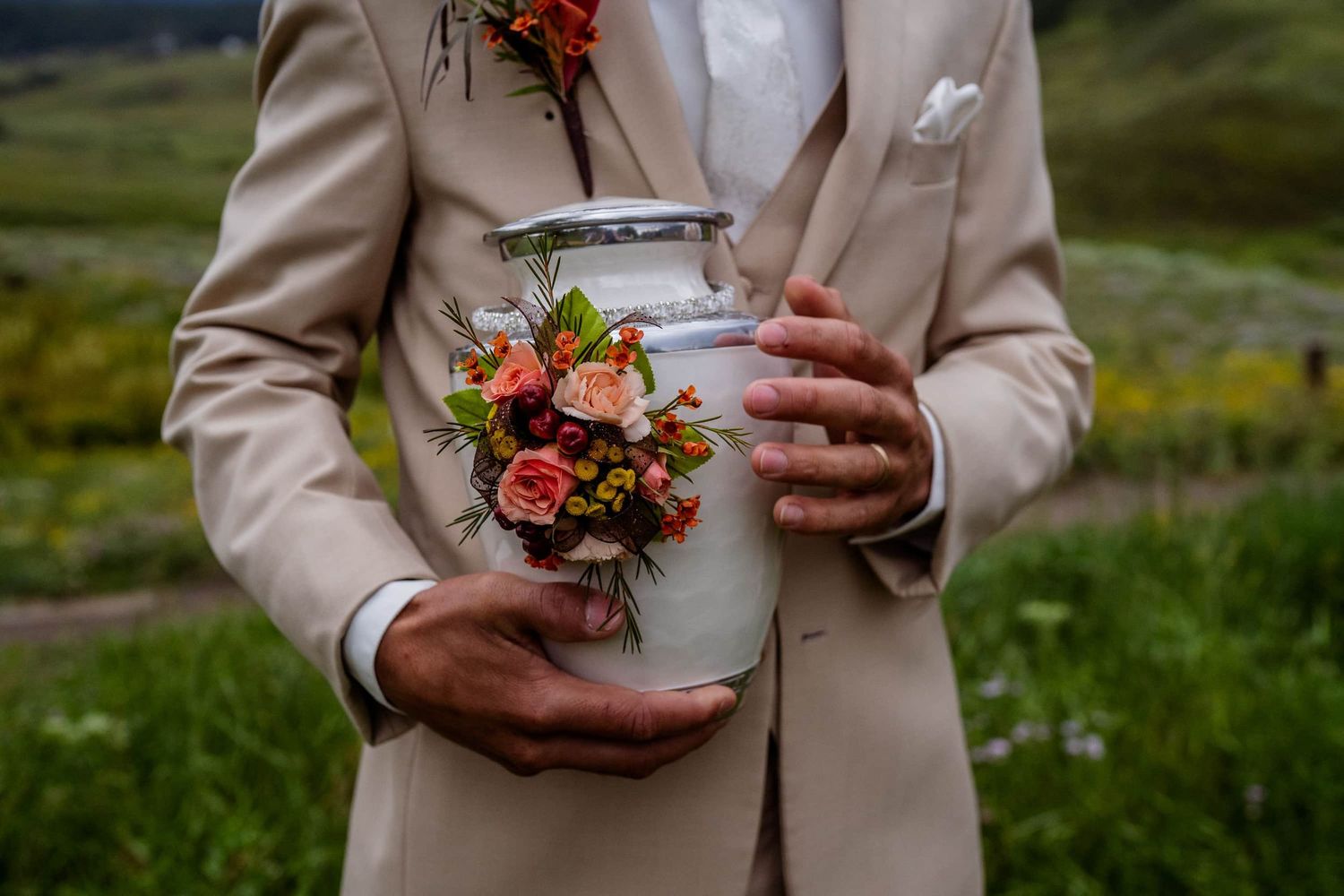 Groom in beige suit holds a decorative white urn with coral floral bouquet against green meadow background.