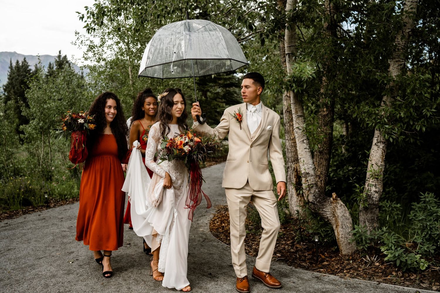 A wedding party walks along a mountain path while someone holds an umbrella over them on an overcast day.